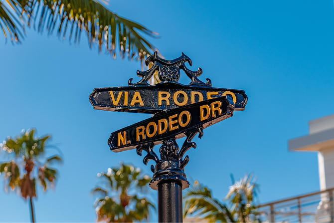 Ornate black and gold street sign for Via Rodeo and N. Rodeo Dr against a clear blue sky with palm trees.