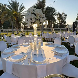 Elegant wedding reception table with white linens, tall floral centerpiece, and palm trees in background.