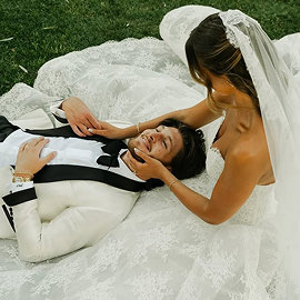 Bride in lace gown and veil lying on grass gently holding groom's face, both smiling.