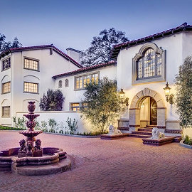 Spanish-style mansion at dusk with illuminated arched entryway, tiled fountain, and terracotta courtyard.