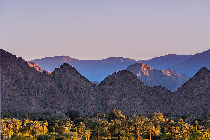 Desert mountain range at sunset with palm trees in the foreground, likely near Palm Springs or La Quinta.
