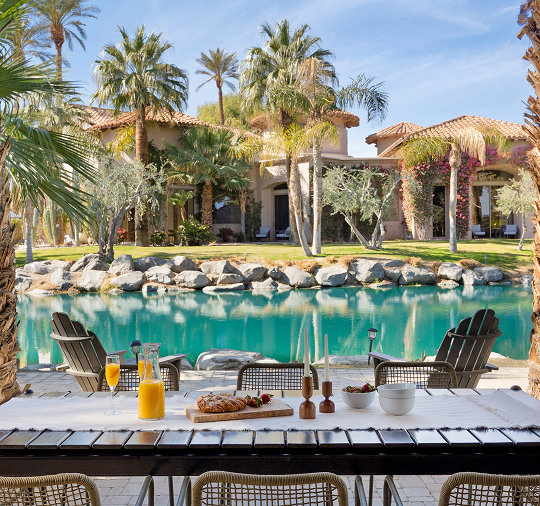 Outdoor dining table set with breakfast pastries and orange juice beside a turquoise lagoon, with a Mediterranean-style estate and palm trees in the background.