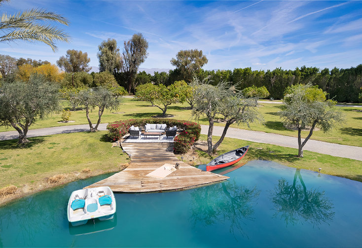 Wooden deck with outdoor seating area extending into a blue pond, surrounded by lush trees and grass, with a paddle boat and canoe docked nearby.