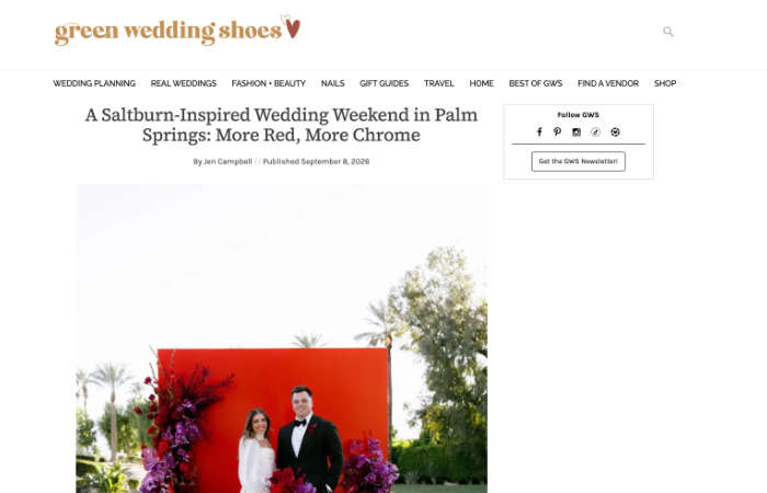 Bride and groom stand in front of a red backdrop adorned with purple flowers, with palm trees and greenery visible in the background.