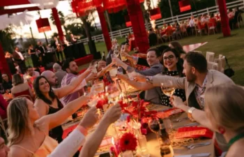 A group of people sit at a long outdoor table decorated with red accents, raising glasses in a toast during an evening celebration.