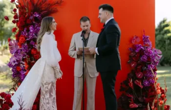 A couple stands facing each other on a red platform decorated with red and purple flowers, with an officiant between them, outdoors under palm trees.