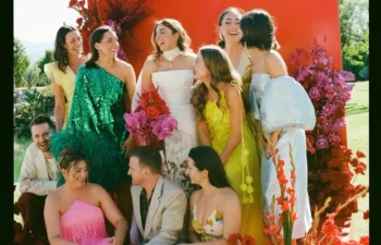 A group of people in colorful formal attire pose and smile in front of a red backdrop with vibrant red and pink flowers outdoors.