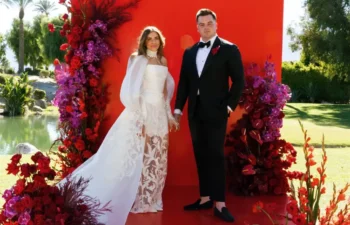 A couple in formal wedding attire stands on a red platform decorated with red and purple flowers, outdoors with palm trees and water in the background.