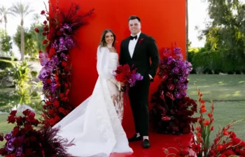 A bride and groom stand on a red platform decorated with red and purple flowers, with trees and a lawn in the background.