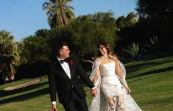 A bride and groom walk hand in hand on a green lawn, surrounded by trees and palm trees, under a clear blue sky.