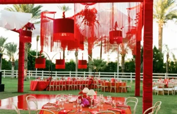 Outdoor event setup with round tables covered in orange and red linens, wooden chairs, and a red-framed canopy with hanging decorations on a grassy lawn.