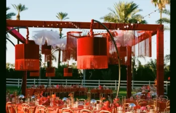 Outdoor event setup with long tables covered in red cloth, matching chairs, red hanging lamps, and palm trees in the background under a clear sky.