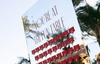 A mirrored seating chart sign outdoors reads “You’re at the fun table” above rows of small red envelopes, with palm trees and buildings visible in the background.