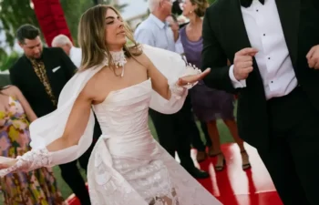A bride in a white dress and a man in a tuxedo dance on a red floor, surrounded by people at an outdoor event.