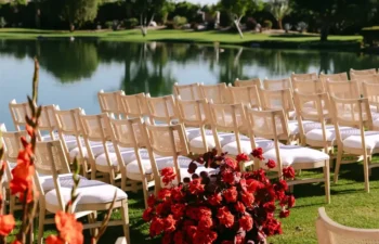 Rows of wooden chairs face a lake, with red floral arrangements on the grass, set outdoors on a sunny day with trees in the background.