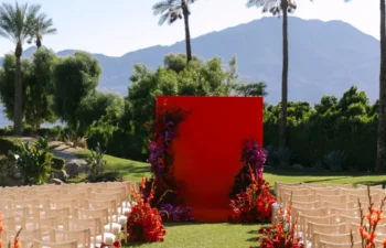 Outdoor wedding setup with rows of chairs, vibrant red floral arrangements, and a large red backdrop, with palm trees and mountains in the background under a clear sky.
