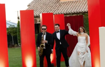 A bride and groom walk on grass holding hands, smiling, followed by a saxophonist. Red vertical panels and a building are in the background.